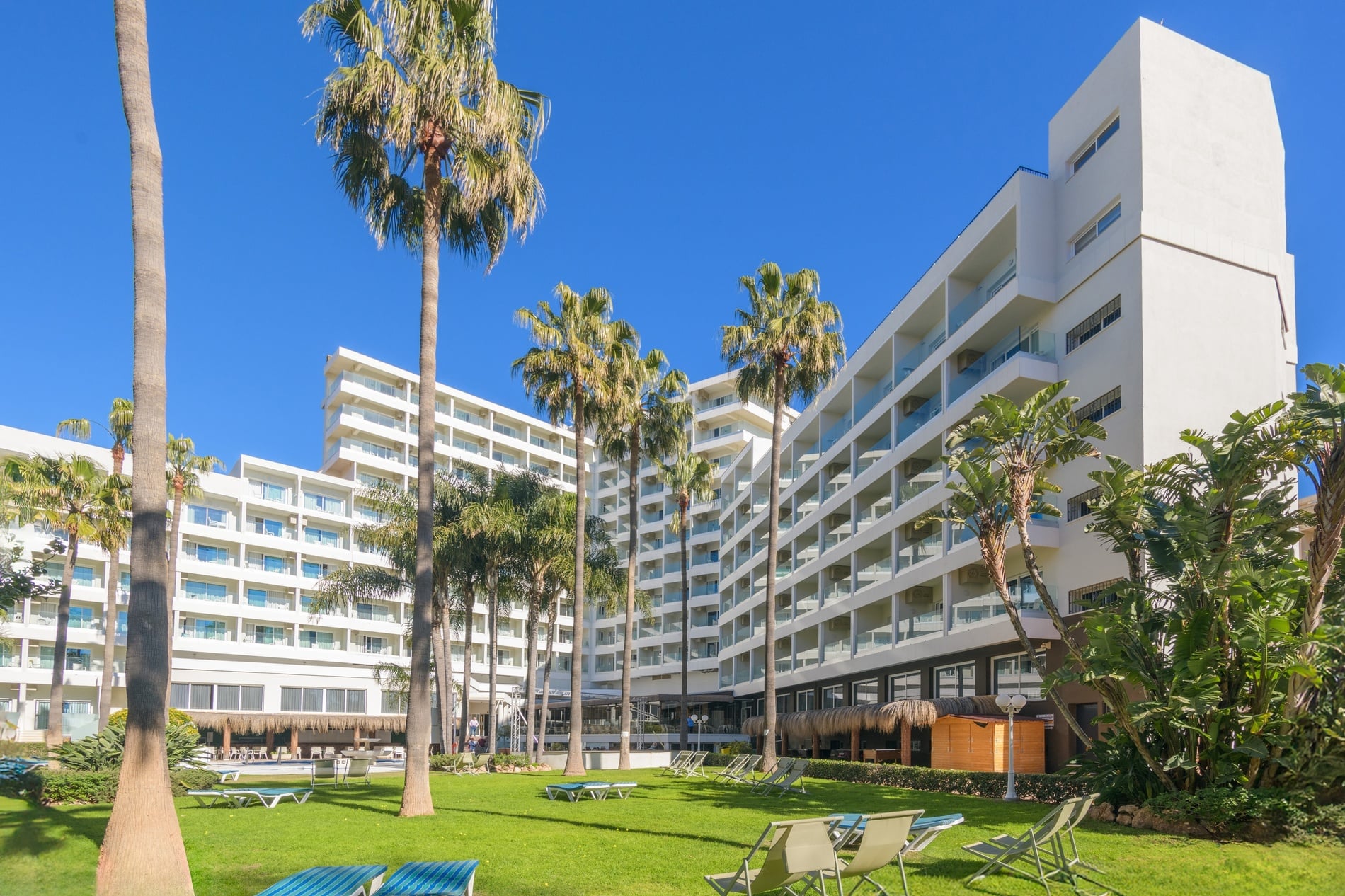 Un hotel moderno y blanco se alza majestuosamente, rodeado de esbeltas palmeras y un exuberante césped verde con tumbonas, todo bajo un cielo azul resplandeciente.