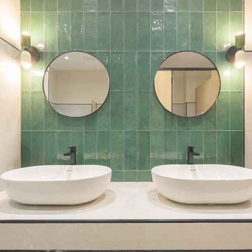 Un baño moderno presenta dos lavabos blancos sobre un mostrador claro, enmarcado por una pared de azulejos verdes, con dos espejos redondos y luces montadas a los lados.