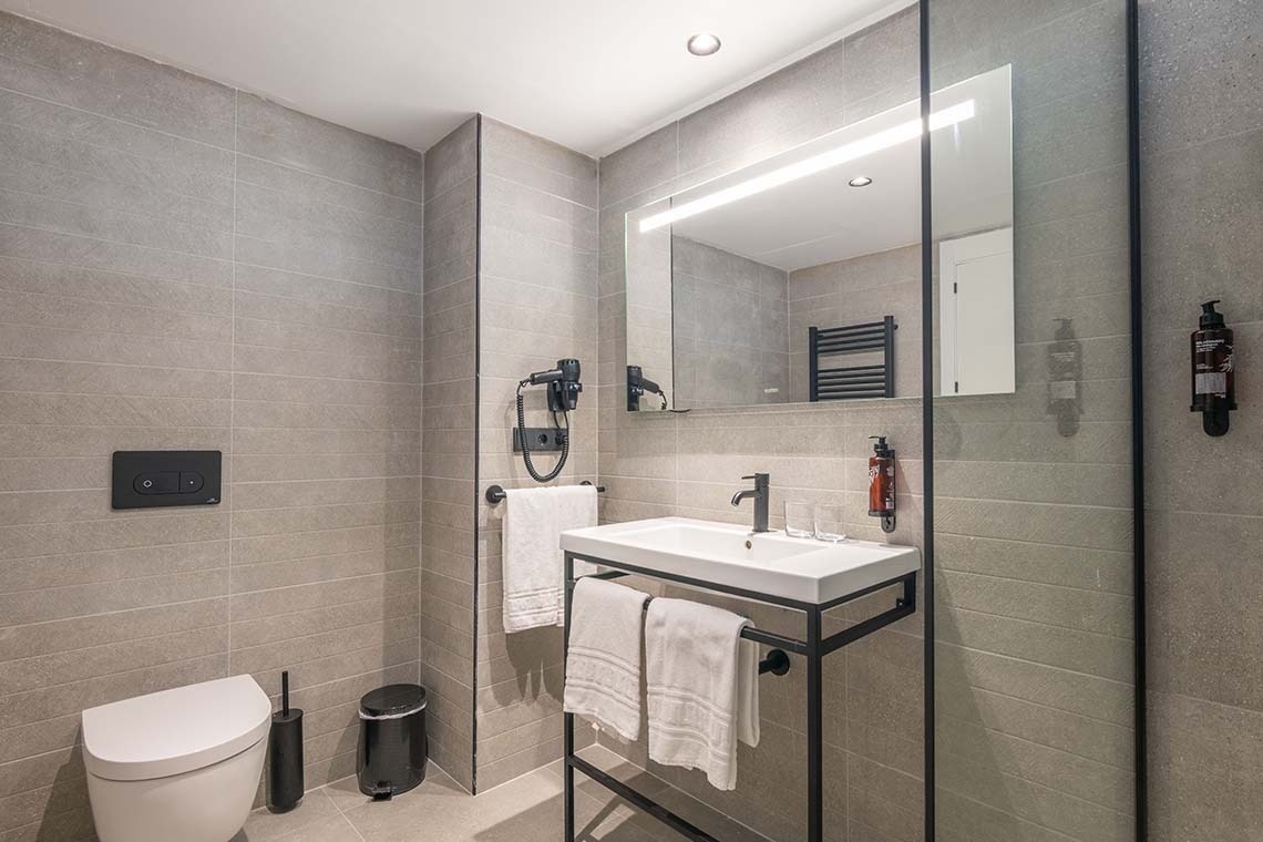 Un baño moderno y funcional presenta paredes de azulejos grises, un inodoro, un lavabo con una elegante estructura metálica negra, un espejo iluminado y varios accesorios en tonos negros.