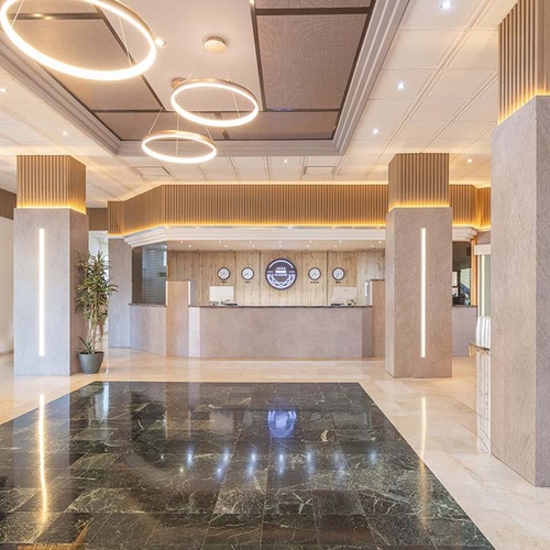 Un moderno lobby de hotel con una recepción central, columnas con iluminación vertical, un suelo de mármol pulido en contraste y llamativas lámparas circulares colgantes.