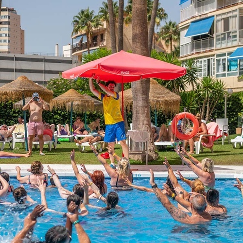 Un grupo de personas participa en ejercicios de aeróbic acuático en una piscina, mientras un monitor dirige la actividad bajo un parasol y otros veraneantes se relajan al sol.