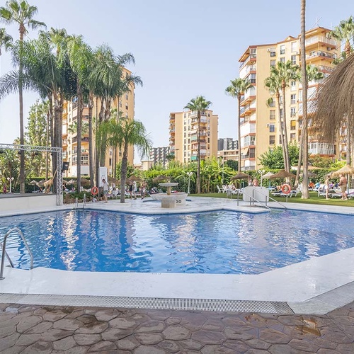 Una gran piscina exterior en un complejo turístico rodeada de palmeras y edificios altos, con personas disfrutando del día soleado.