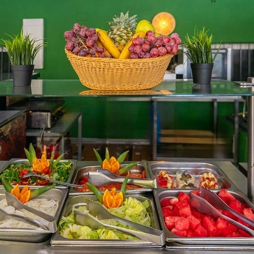 Una línea de buffet vibrante exhibe una variedad de ensaladas, sandía y una cesta de frutas frescas sobre una encimera de cristal en un entorno de pared verde.
