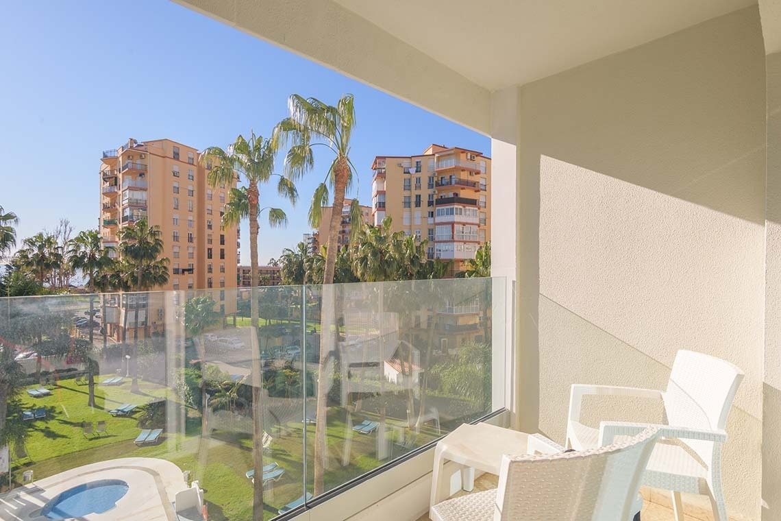Balcón soleado con barandilla de cristal, ofreciendo vistas a un jardín tropical con palmeras, edificios y una pequeña piscina, con mobiliario exterior blanco.