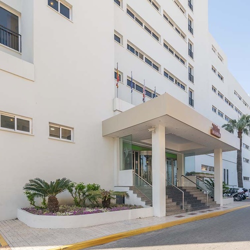 La fachada de un moderno edificio blanco, posiblemente un hotel, se muestra con su entrada principal cubierta, escaleras y paisajismo con palmeras, bajo un cielo despejado.
