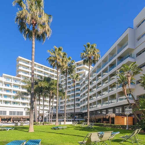 Un moderno complejo hotelero blanco con múltiples edificios y balcones se extiende entre palmeras altas y césped verde, bajo un cielo azul brillante.