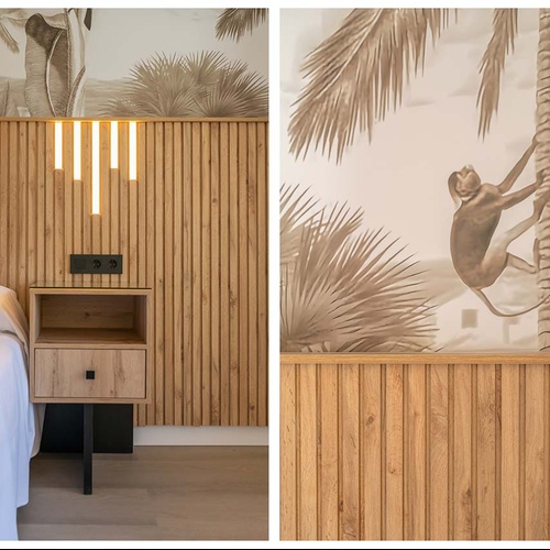 Una habitación moderna con un cabecero de lamas de madera, iluminación vertical, una mesita de noche y un mural decorativo tropical que incluye palmeras y un mono.
