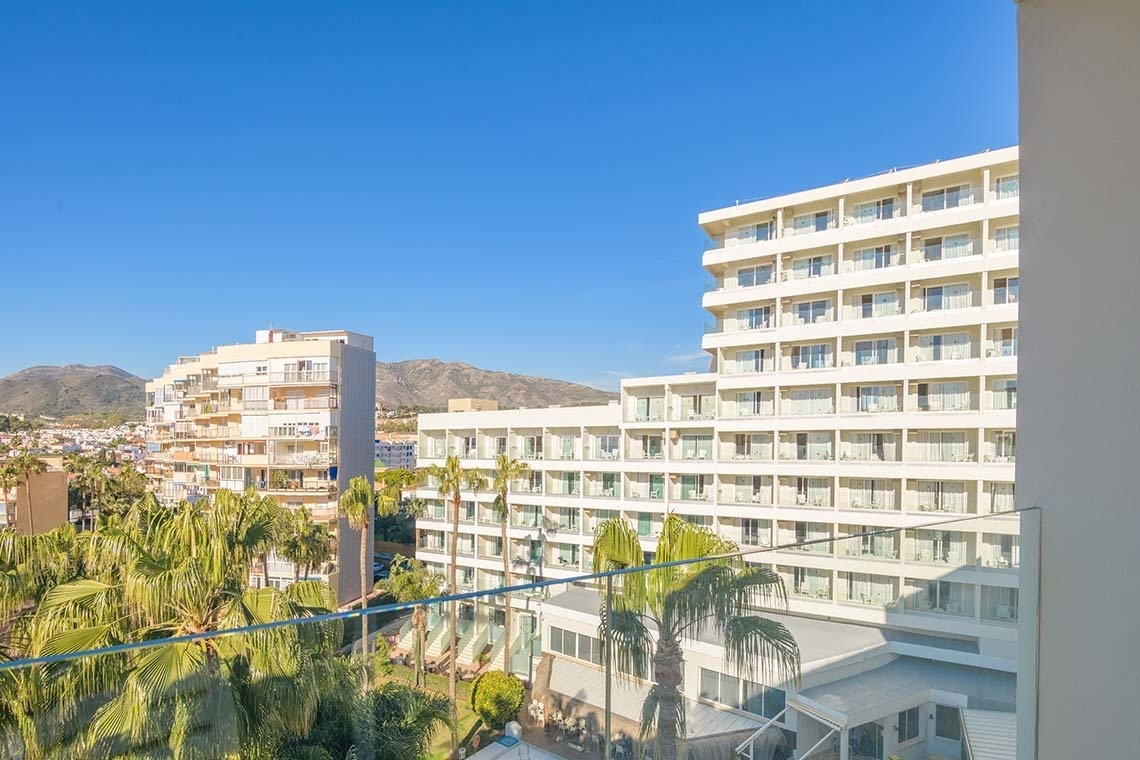 Vista desde un balcón de un complejo de edificios blancos y palmeras, con montañas en la distancia bajo un cielo azul claro.