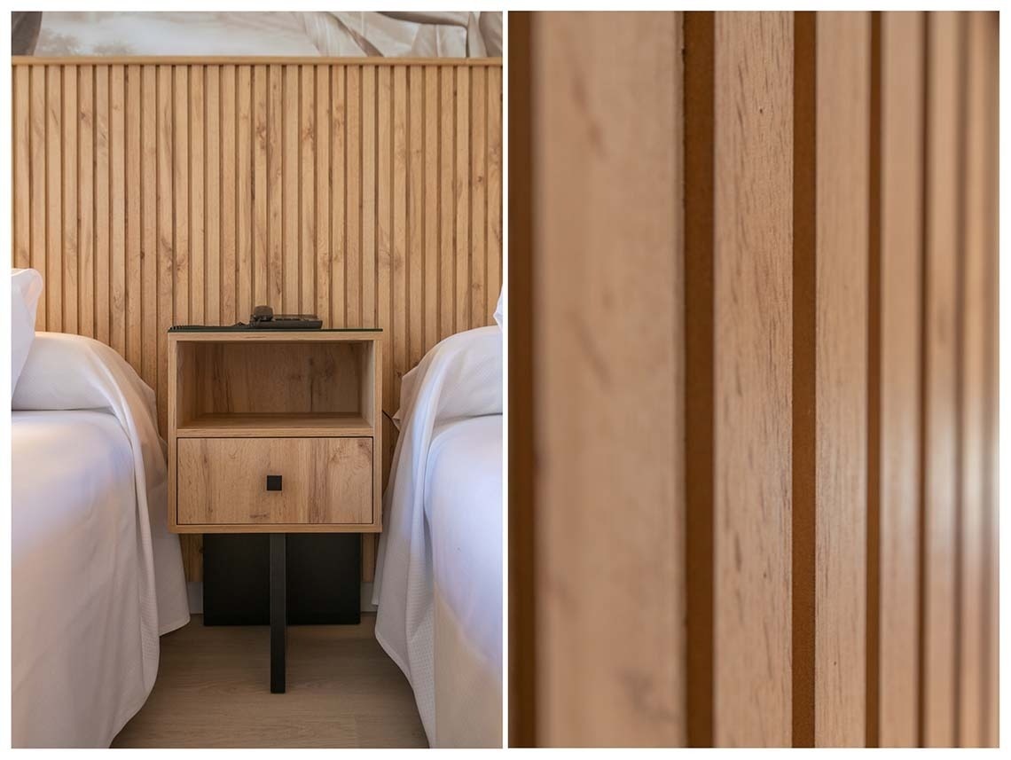 Una mesita de noche de madera se encuentra entre dos camas, respaldada por un cabecero de lamas de madera, cuyo detalle se aprecia en un primer plano.