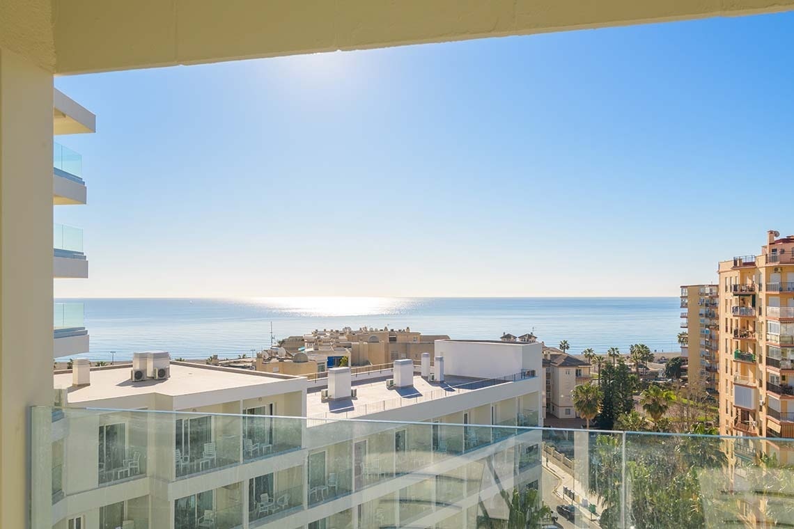 Desde un balcón, se observa un paisaje costero soleado con el mar azul brillante, edificios modernos y palmeras.