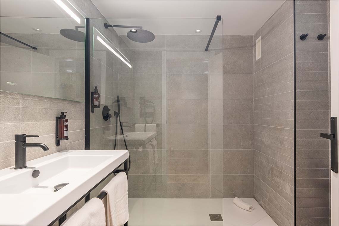Un moderno cuarto de baño minimalista presenta una ducha con mampara de cristal, azulejos grises, y un lavabo blanco con accesorios negros.