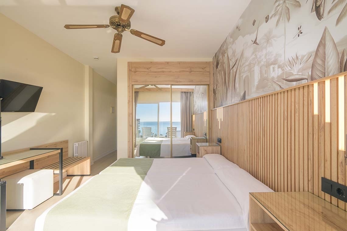 Un luminoso dormitorio de hotel de estilo moderno, con un mural tropical y un ventilador de techo, que ofrece una vista al mar reflejada en las puertas del armario.