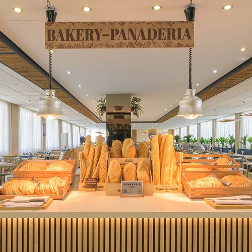 Una sección de panadería con un mostrador que exhibe varios tipos de pan fresco y un letrero de 