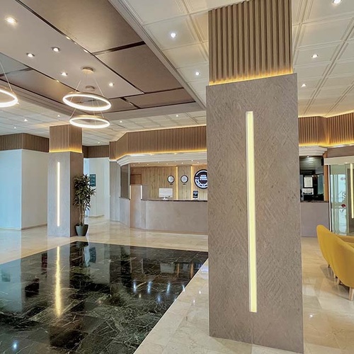 Luminoso vestíbulo de hotel moderno con suelo de mármol pulido, pilares decorados con madera e iluminación integrada, y un mostrador de recepción al fondo.