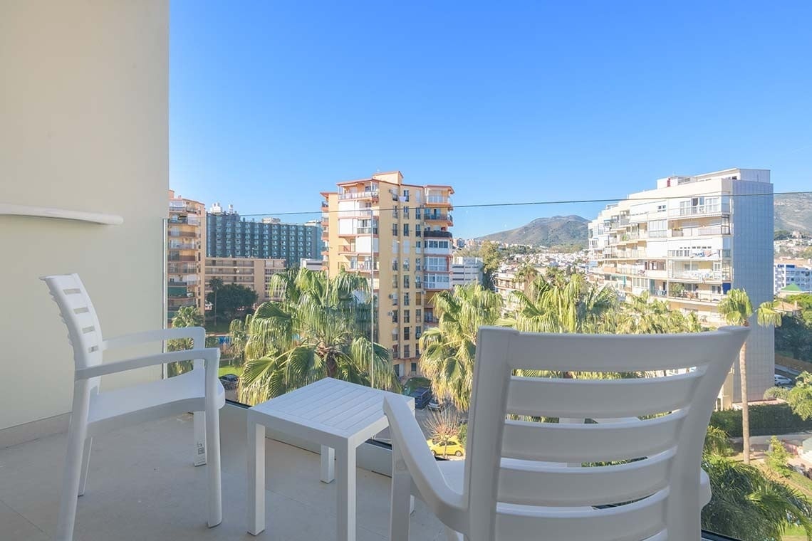 Una vista desde un balcón con muebles blancos revela una ciudad con edificios, palmeras y montañas en el horizonte bajo un cielo azul despejado.