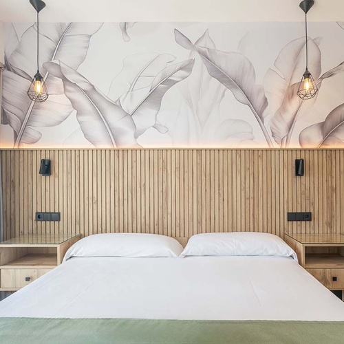 Un dormitorio moderno con una cama doble, un cabecero de madera, papel tapiz de hojas de palma, dos lámparas colgantes y mesitas de noche a cada lado.