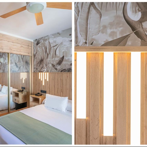 Una moderna habitación de hotel presenta una cama con cabecero de madera y luces LED, papel pintado tropical en tonos sepia, un armario con espejo y un ventilador de techo.