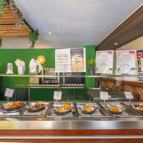 Un bar de snacks ofrece una variedad de comida caliente en bandejas, con carteles informativos sobre el menú y el horario, todo dentro de un interior con paredes verdes.
