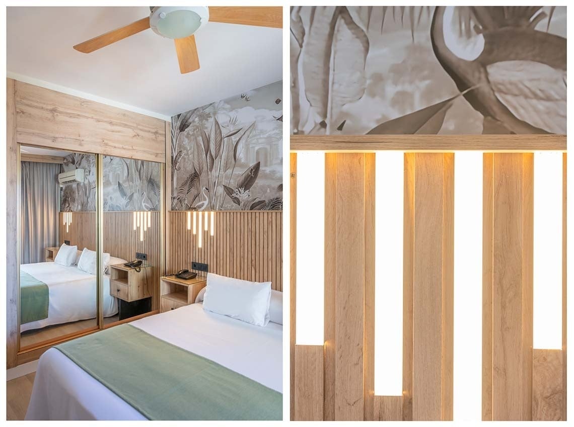 La imagen muestra una habitación de hotel con un cabecero de lamas de madera iluminadas, un mural tropical en tonos sepia, un armario con puertas de espejo y un ventilador de techo.