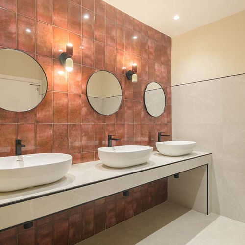 Un moderno baño presenta tres lavabos con sus respectivos espejos redondos, destacando una pared de azulejos en tonos rojizos.