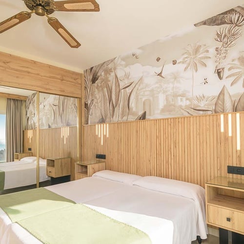 Habitación de hotel con decoración tropical que presenta un cabecero de madera con luces integradas, papel pintado con motivos de la jungla y un armario con espejos que reflejan el mar.