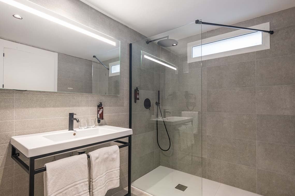 Un baño moderno con paredes de azulejos grises, un lavabo blanco con grifo negro y un gran espejo iluminado sobre él, junto a una ducha a ras de suelo con mampara de cristal y detalles en negro.