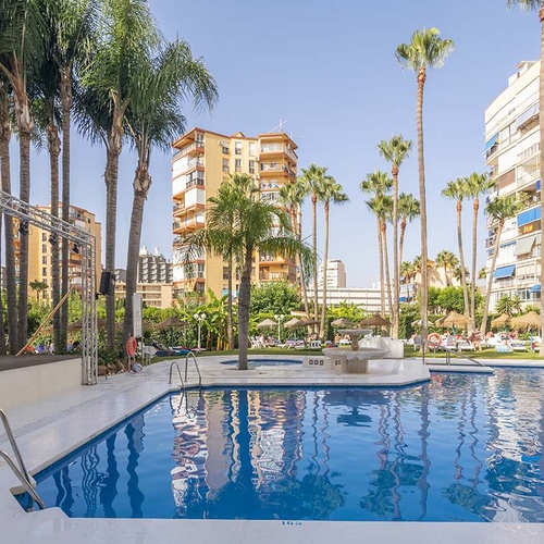 Una piscina al aire libre rodeada de palmeras y edificios altos bajo un cielo azul despejado.