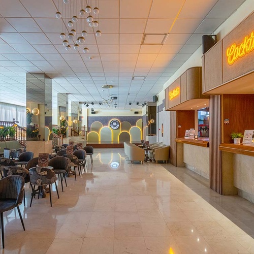 Una amplia y moderna zona de bar/salón de hotel con asientos, una barra de cócteles iluminada y un escenario al fondo.