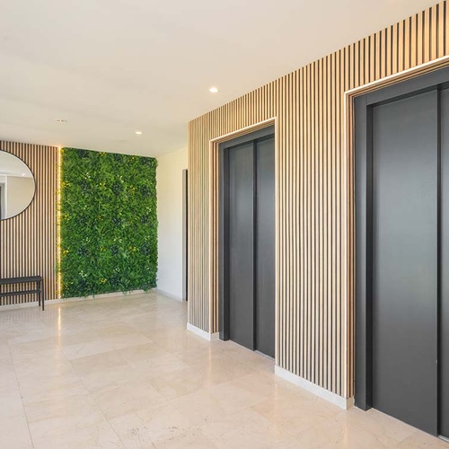 Un moderno vestíbulo de un edificio presenta dos ascensores grises, paredes decoradas con listones de madera y un jardín vertical, complementado con un espejo redondo y un banco.