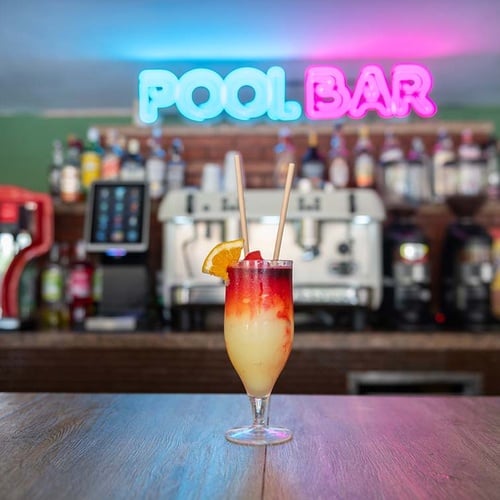 Un colorido cóctel con una rodaja de naranja y pajitas se exhibe en una barra de madera, con un letrero de neón 