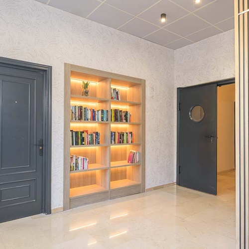 Un pasillo interior presenta dos puertas oscuras, una estantería empotrada de madera iluminada con libros, paredes con papel tapiz de hojas claras y un panel decorativo de listones de madera en el lado derecho.