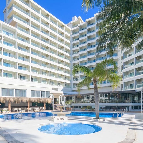 Un complejo hotelero blanco y moderno con varias piscinas y palmeras se extiende bajo un cielo azul despejado.