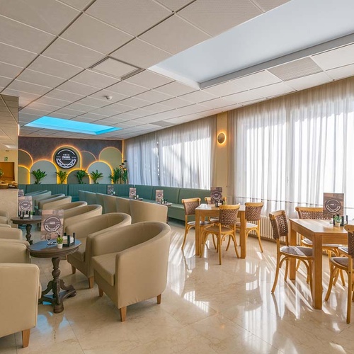 Un amplio y luminoso salón de hotel o cafetería ofrece diversas zonas de asiento con mobiliario moderno y clásicos sillones, iluminado por grandes ventanales con cortinas blancas.