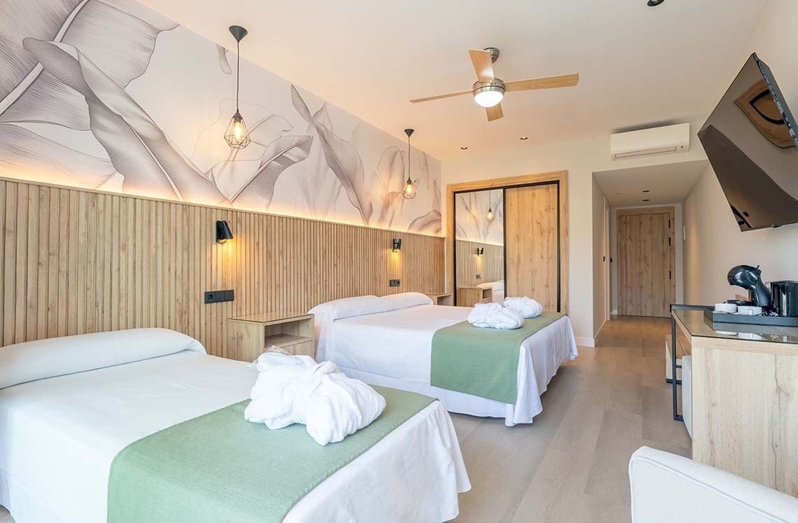 La imagen muestra una moderna habitación de hotel con dos camas, una cabecera de madera y papel tapiz botánico, equipada con ventilador de techo, TV y un área de café.