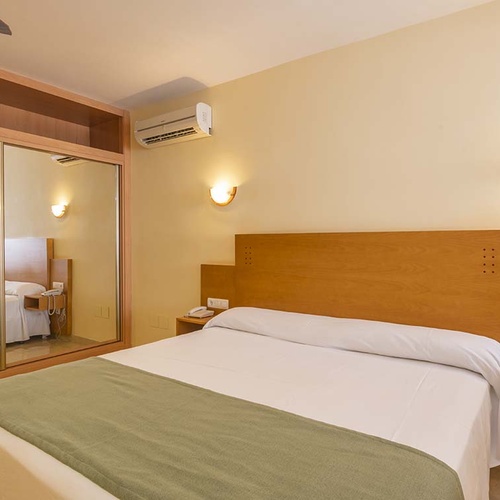 Un dormitorio luminoso con cama doble, cabecero de madera, mesitas de noche, un armario con espejo que refleja otra cama y una ventana de pavés, paredes crema, aire acondicionado y ventilador de techo.