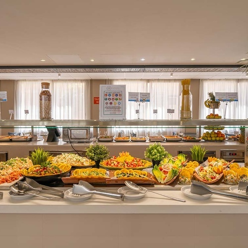 Una mesa de buffet generosamente surtida exhibe una variedad de ensaladas, platos preparados y aderezos en un entorno luminoso y acogedor.