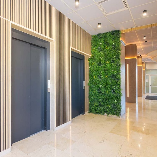 Un moderno vestíbulo presenta dos ascensores con puertas oscuras, paredes de listones de madera clara, una gran pared vegetal y un reluciente suelo de mármol.