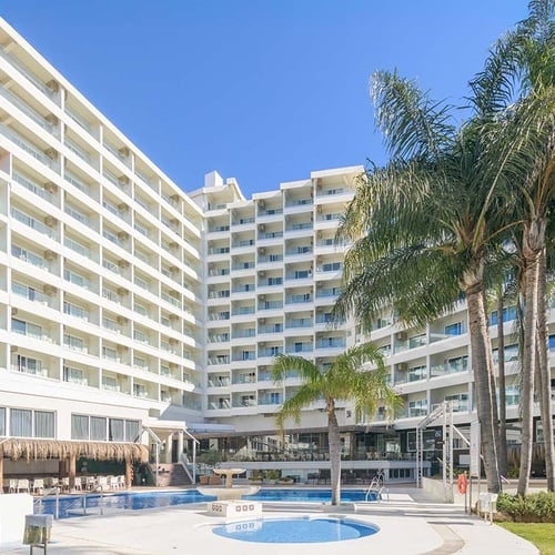 Un gran complejo hotelero blanco en forma de U, con múltiples balcones y palmeras, rodea una zona de piscinas al aire libre bajo un cielo azul claro.