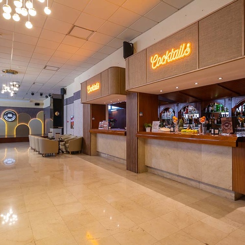 La imagen muestra un amplio bar o lounge interior con un letrero de neón que dice 