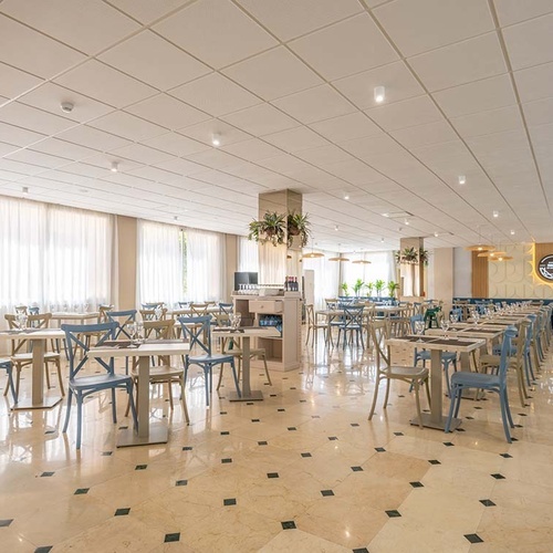 Un amplio y luminoso comedor de restaurante se presenta con numerosas mesas y sillas de tonos claros, suelo de mármol decorado con cuadros oscuros y grandes ventanales con cortinas blancas, bajo un techo de paneles acústicos.