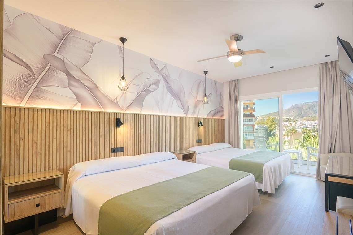 Una habitación de hotel moderna y luminosa con dos camas, un cabecero de madera con lamas, papel pintado de hojas tropicales y un balcón con vistas a la ciudad y las montañas.