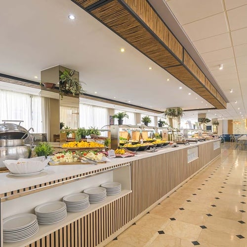 Un amplio y luminoso comedor de hotel con una extensa barra de buffet repleta de alimentos y mesas y sillas preparadas para los comensales.