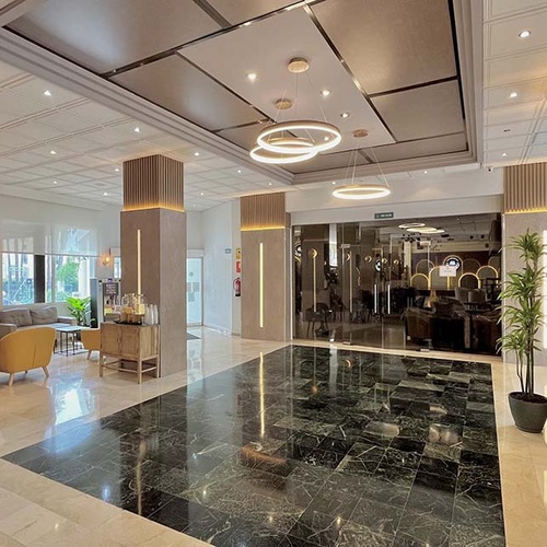 Un moderno y elegante vestíbulo de hotel se presenta con un llamativo suelo de mármol pulido bicolor, columnas iluminadas y lámparas colgantes circulares, creando un ambiente sofisticado.