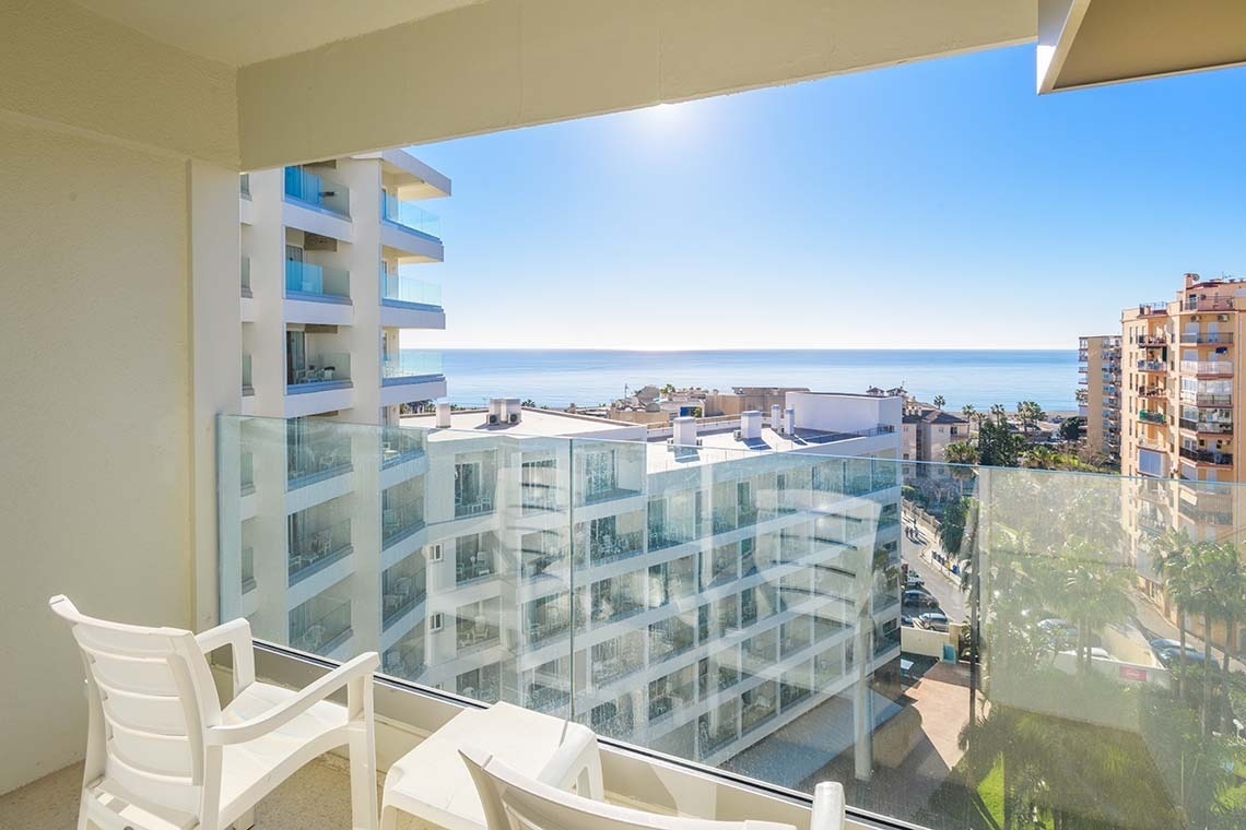 Vista luminosa desde un balcón con sillas blancas hacia el mar azul y edificios en un día soleado.