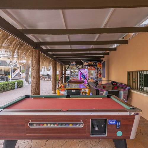 Una zona de juegos al aire libre con una mesa de billar, mesas de air hockey y otras máquinas recreativas bajo una pérgola, con un complejo turístico de fondo.