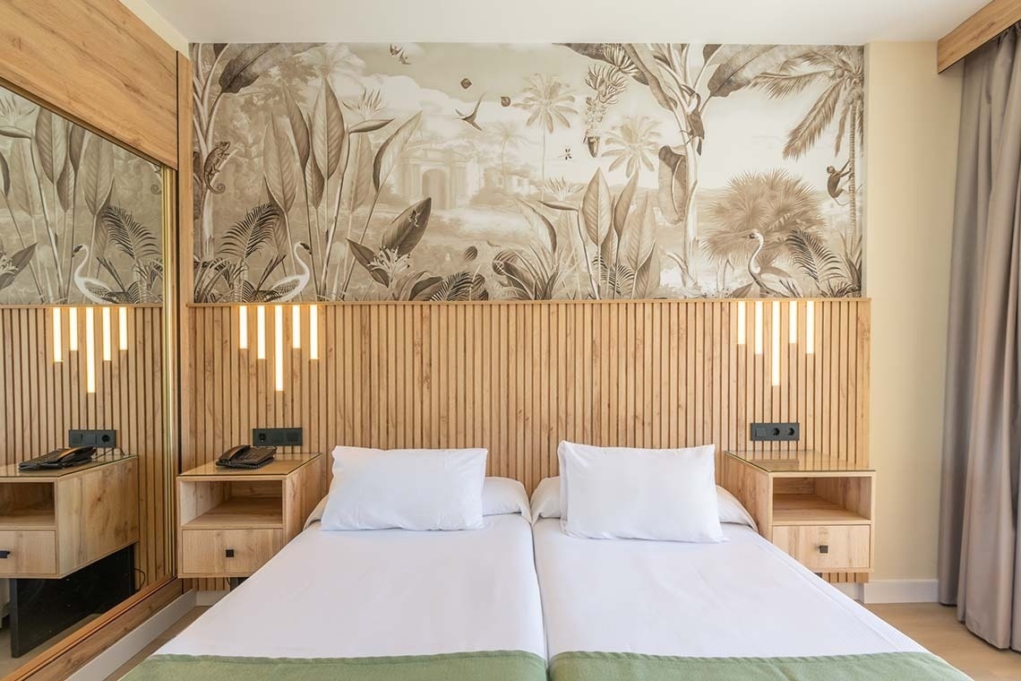 Una habitación de hotel con dos camas individuales presenta un cabecero de madera con lamas y luces verticales modernas, complementado por un papel pintado mural tropical con animales y vegetación.