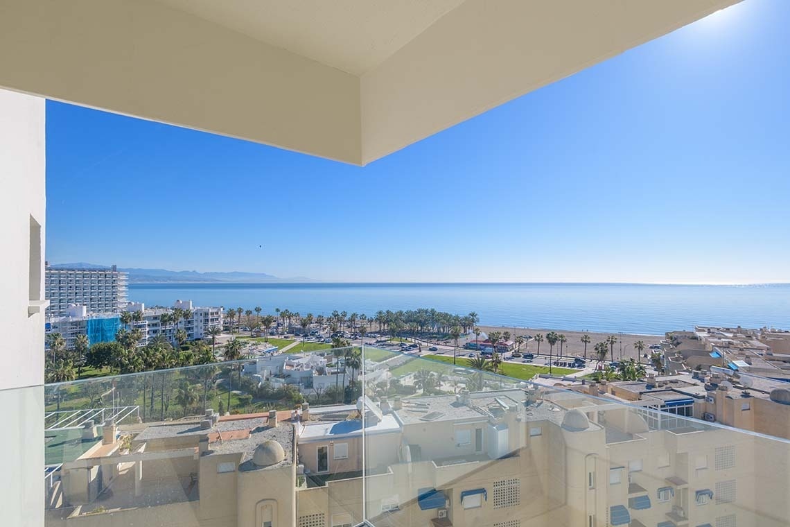 Una vista panorámica desde un balcón de una ciudad costera con un cielo azul despejado, el mar y una playa con palmeras.