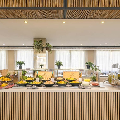 Un variado bufé de ensaladas y platos se exhibe en un comedor luminoso con decoración moderna y natural, destacando un mostrador de madera y un techo de bambú.