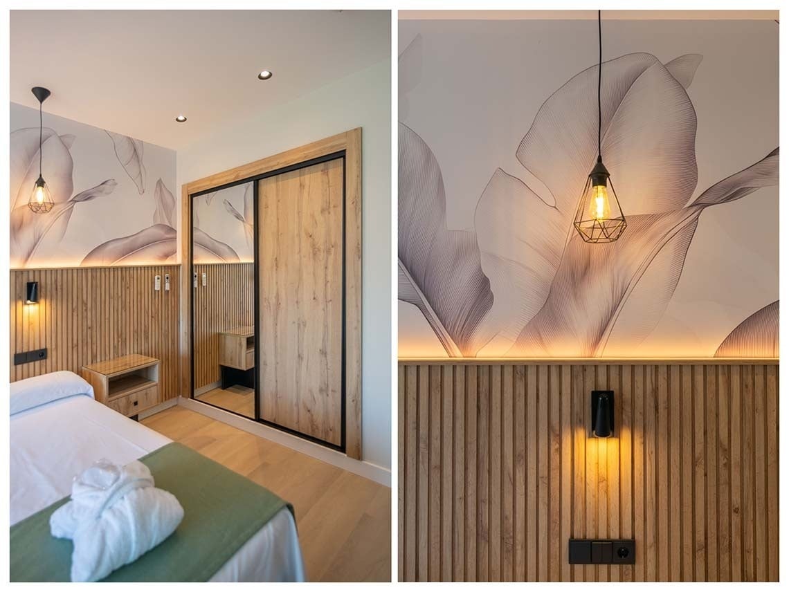 Una habitación moderna y acogedora con un cabecero de madera acanalada, papel pintado de hojas sutiles, un armario de puertas correderas y una iluminación ambiental cálida.