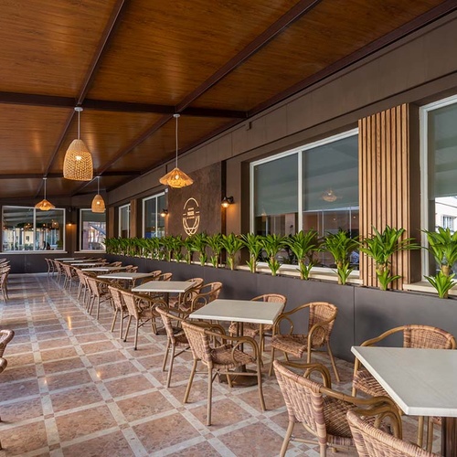 Una terraza de restaurante con mesas y sillas de mimbre bajo un techo de madera con lámparas colgantes y jardineras con plantas a lo largo de las ventanas.